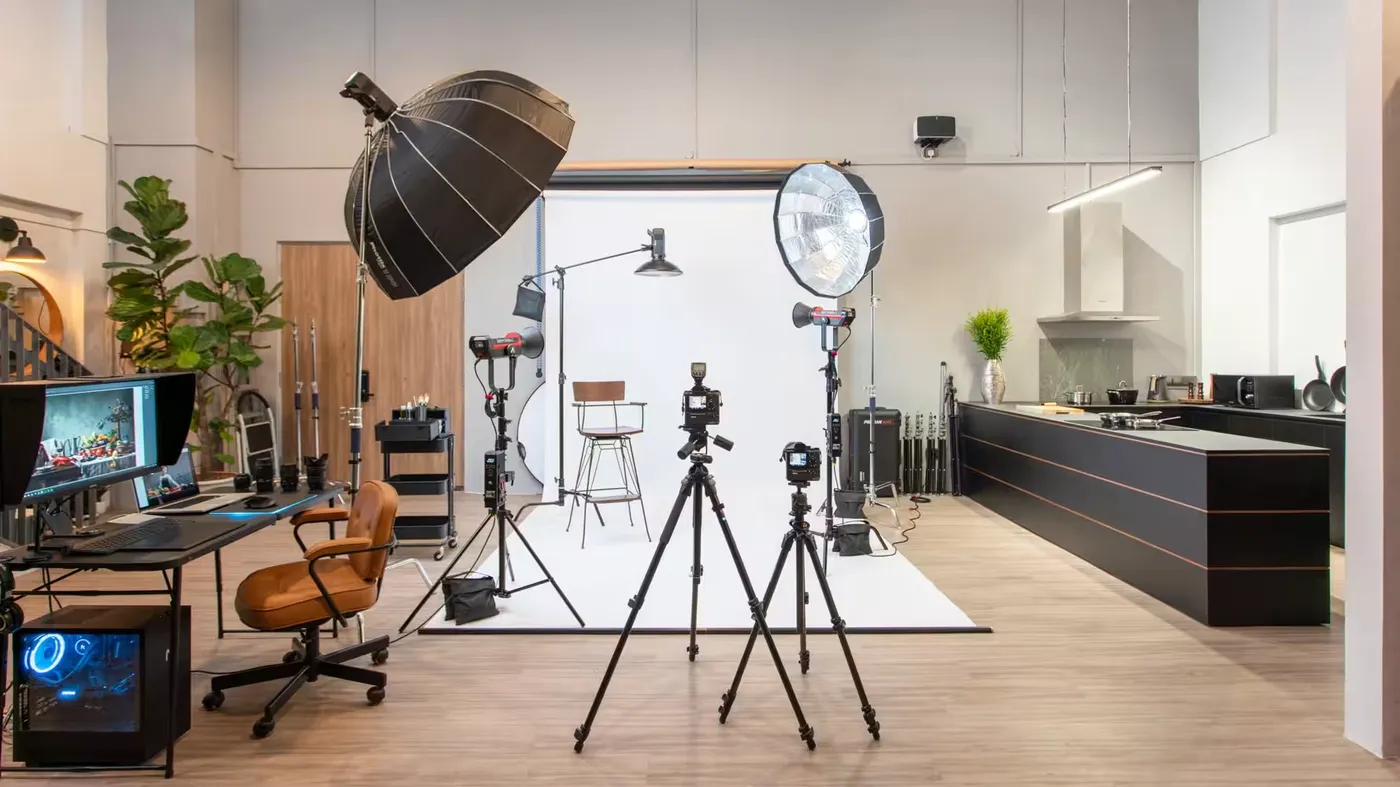 Complete professional photography studio featuring multiple camera setups on tripods facing a white cyclorama backdrop. The versatile space includes professional lighting with a large softbox and beauty dish, an editing workstation with monitor displaying an image, and modern kitchen area. Green plants add warmth to this multi-purpose creative space.