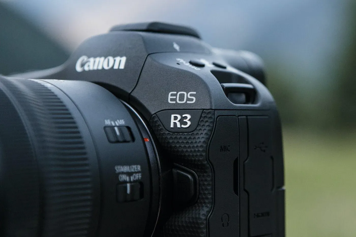 Close-up detail shot of a Canon EOS R3 professional camera body with attached lens. The image clearly shows the camera