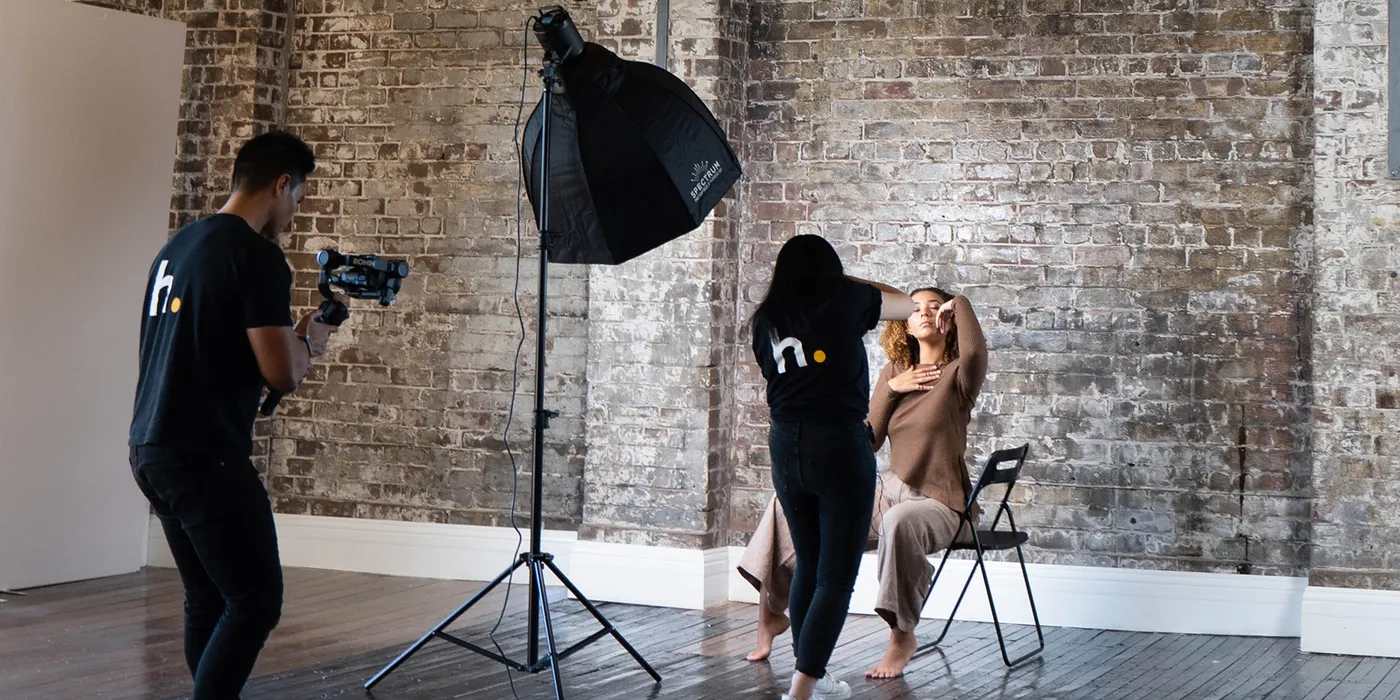 Portrait photography session in an industrial studio with exposed brick walls. Two crew members wearing matching black shirts with logo assist with capturing a model seated on a folding chair. Professional lighting equipment includes a black softbox on a light stand, creating a contrast between the rustic background and controlled lighting.