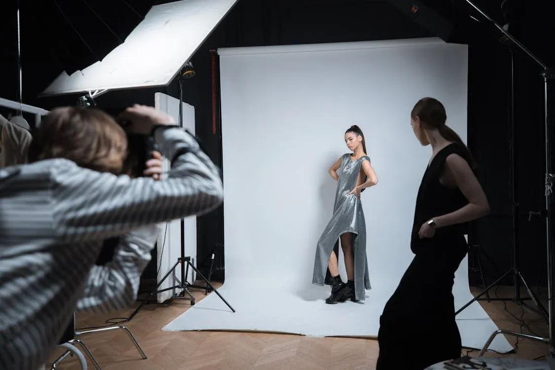 Professional fashion photography session in a studio featuring a photographer capturing a model in a shimmering silver dress against a white backdrop. Studio equipment includes large softbox lighting and a black backdrop setup, with wooden herringbone flooring visible throughout the space.