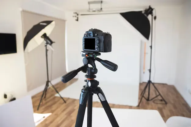 Professional photography studio setup featuring a DSLR camera mounted on a tripod in the foreground with softbox lighting equipment positioned around a white backdrop. The studio has wooden flooring and is arranged for portrait or product photography.