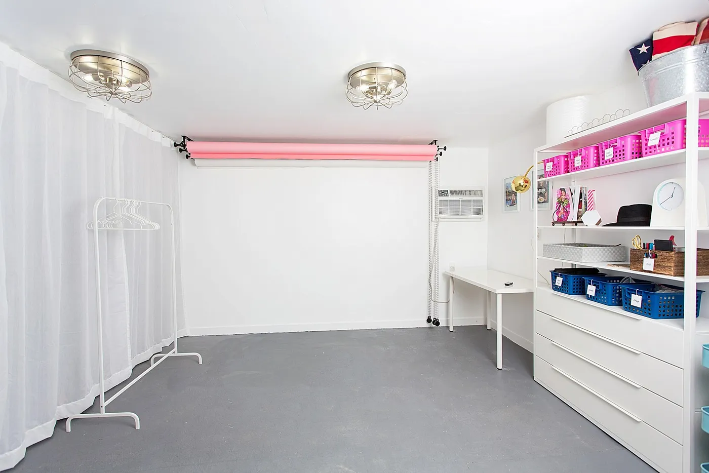 A clean, minimalist home photography studio with white walls and gray flooring. Features include a wall-mounted pink paper backdrop, white curtains for diffusion, a clothing rack, and organized storage shelves with colorful bins. Two industrial-style ceiling lights illuminate the functional workspace.