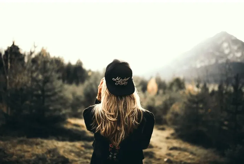 A woman with long blonde hair wearing a black cap and jacket stands with her back to the camera, looking out at a scenic mountain landscape with trees and sunlight.